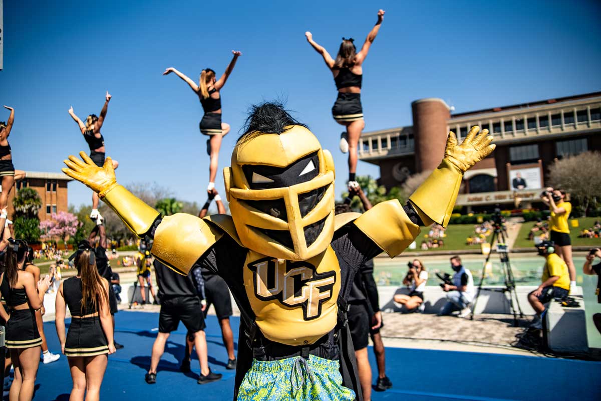 Knightro poses with arms stretched out in front of the cheerleaders stunting