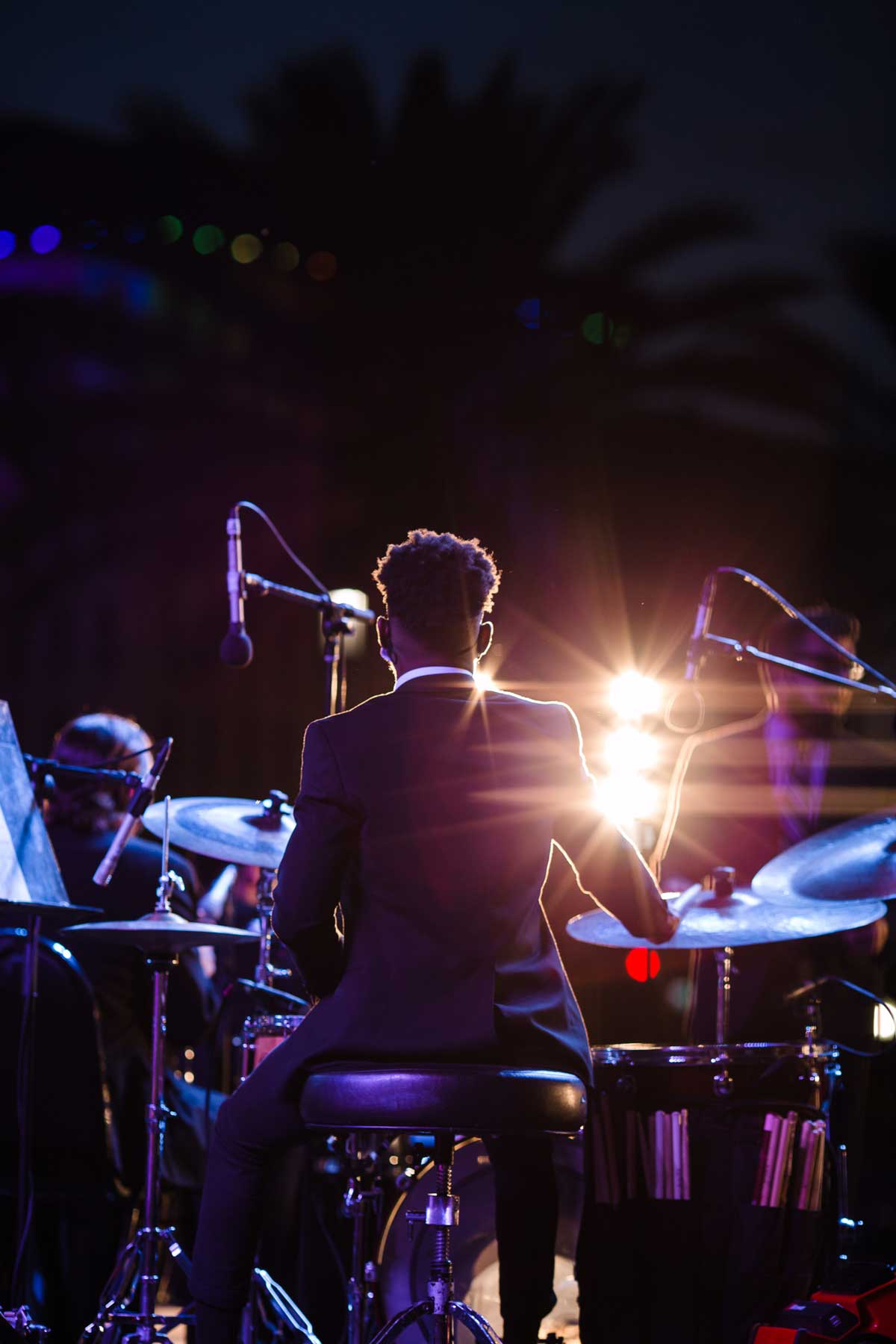 Photo of student seated at drums on stage at night, view from behind