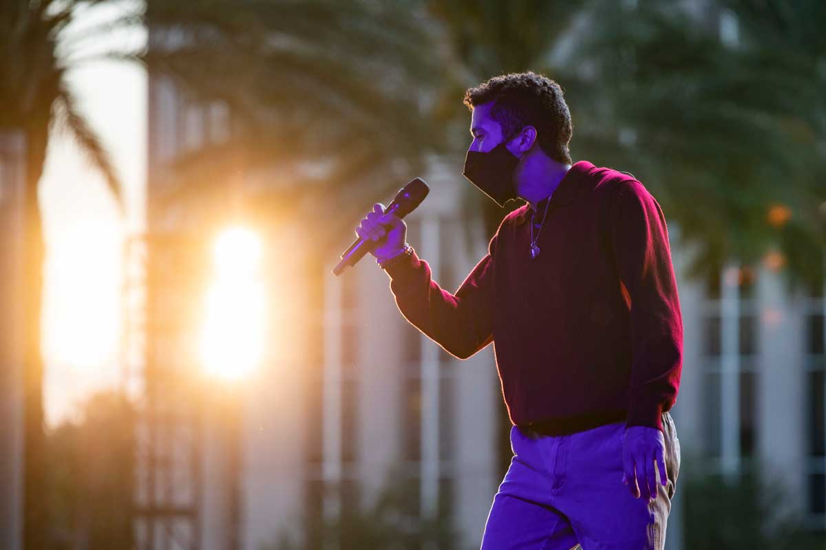 Man sings into a microphone while wearing a mask as the sun sets behind a building in the distance