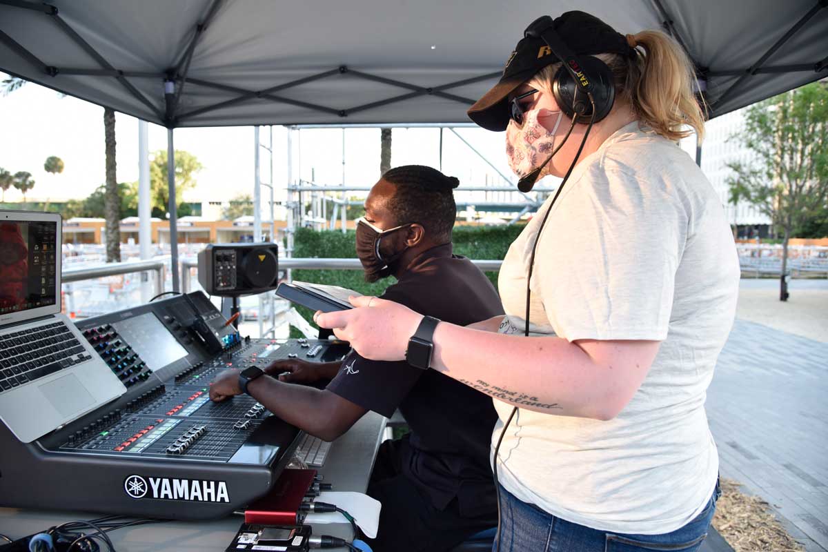 Woman with headset stands next to man seated at sound board under a tent