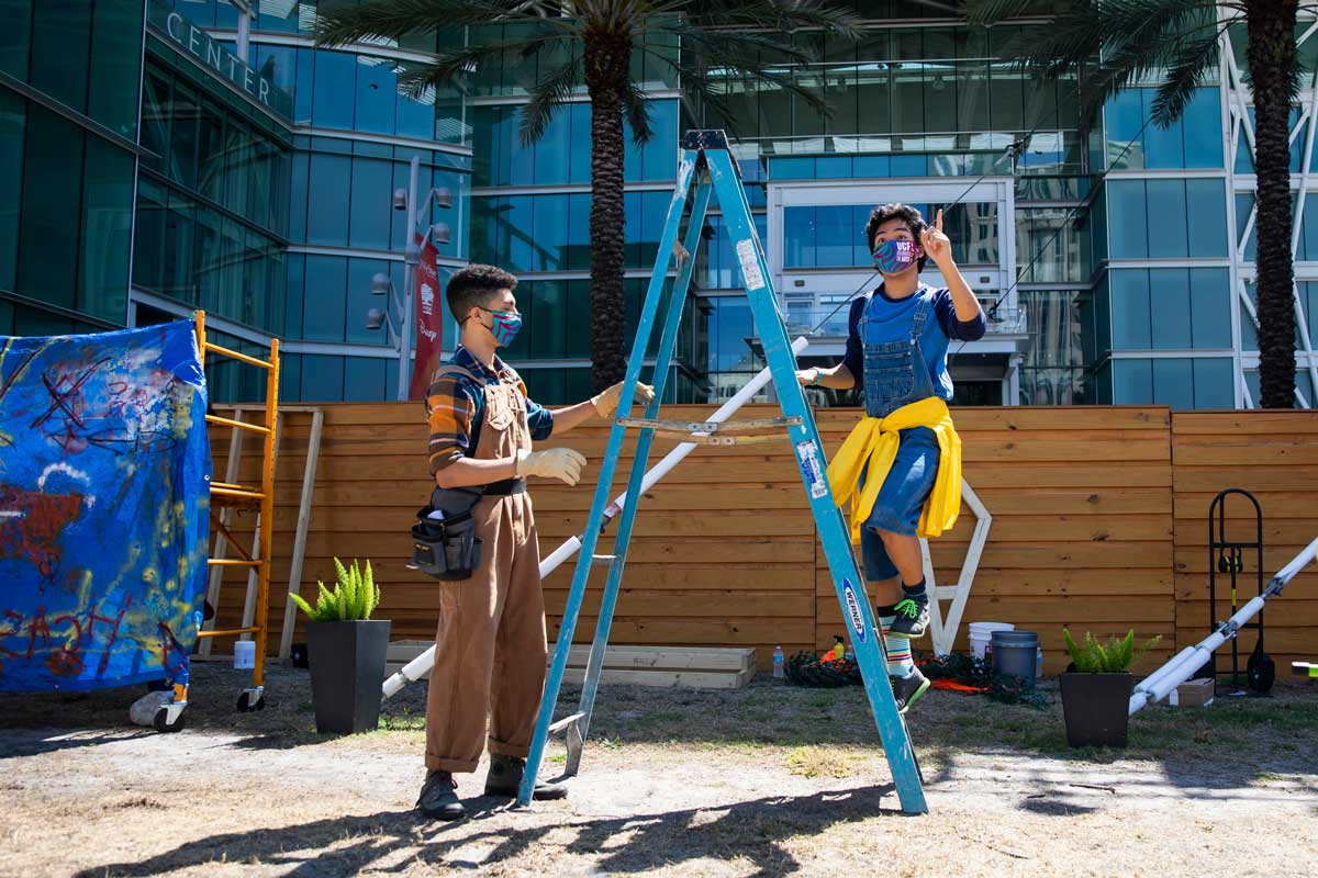 Scene from outdoor production of Grumpiest Boy in the World: Man stands on a ladder dressed as construction worker with another man standing near him