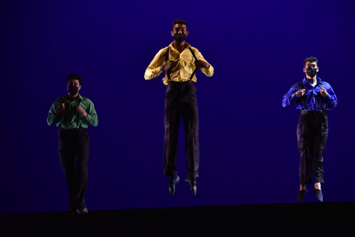 Three dancers wearing dress shirts and pants with suspendersjump on stage 