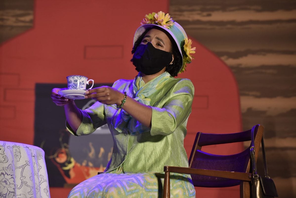 Black woman in costume lift up tea cup and saucer while sitting in a wooden chair on stage
