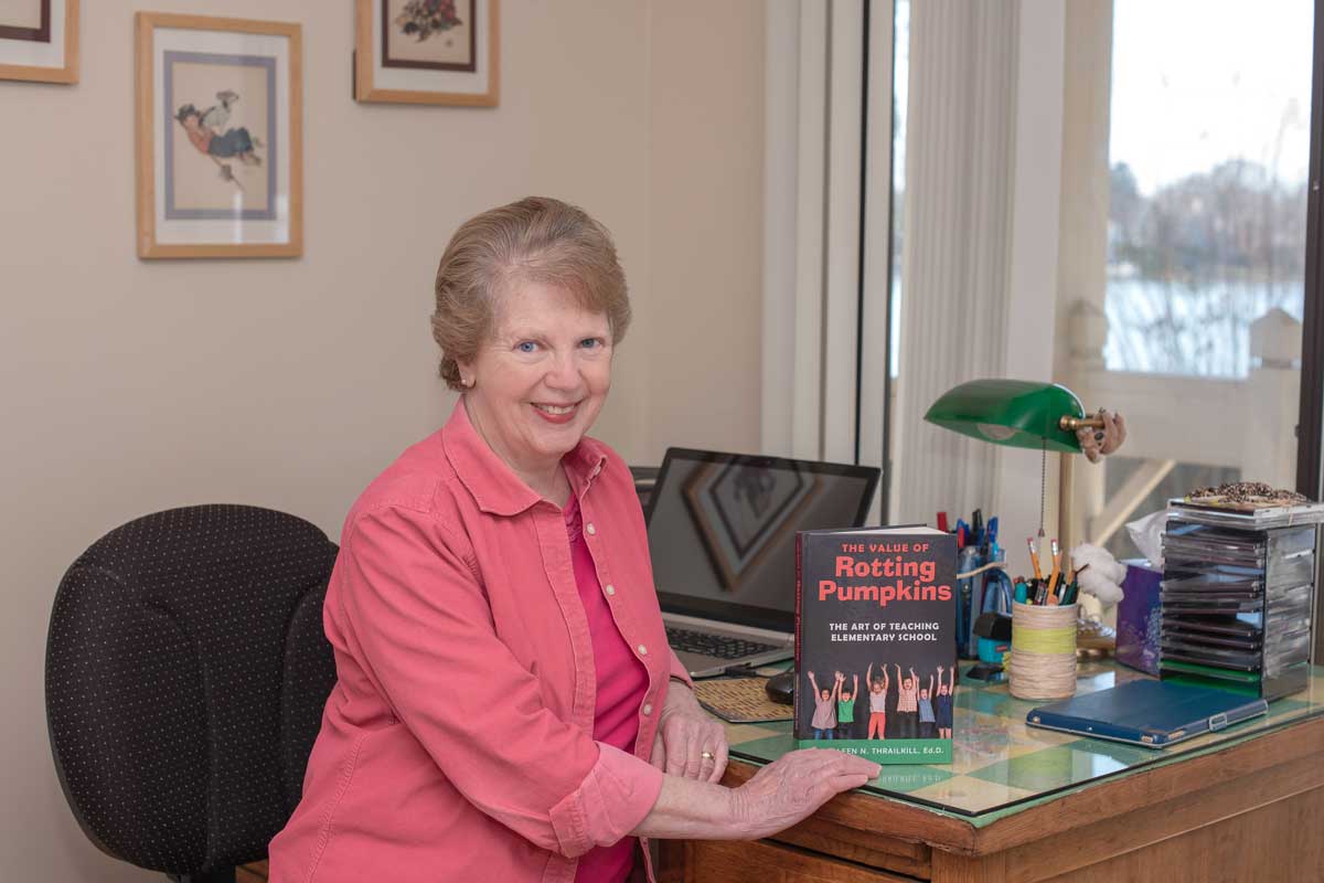 Senior woman wearing pink blouse sits at a desk with a book propped up, The Value of Rotting Pumpkins