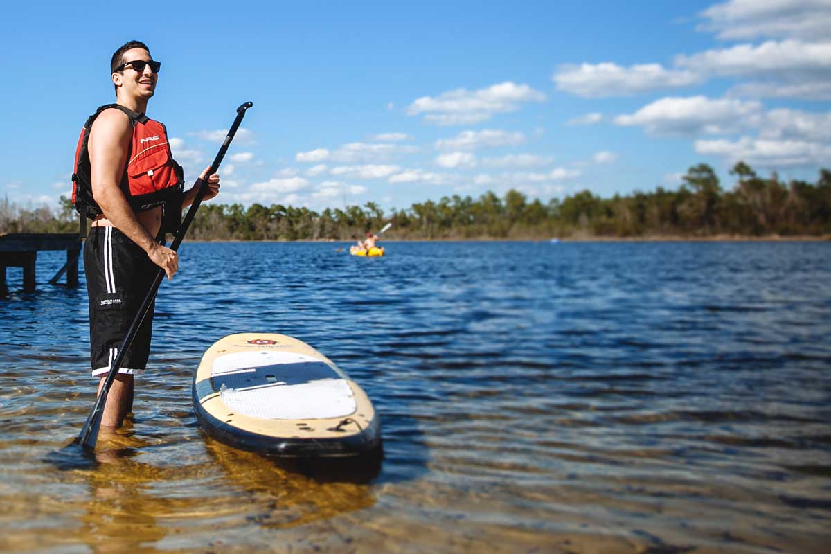 Man in life jacket holds paddle standing next to paddleboard in water