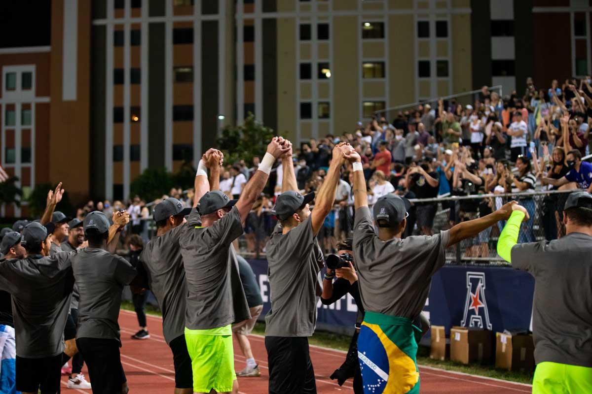 Men's soccer players join hands overhead and face crowd in stands to show thanks