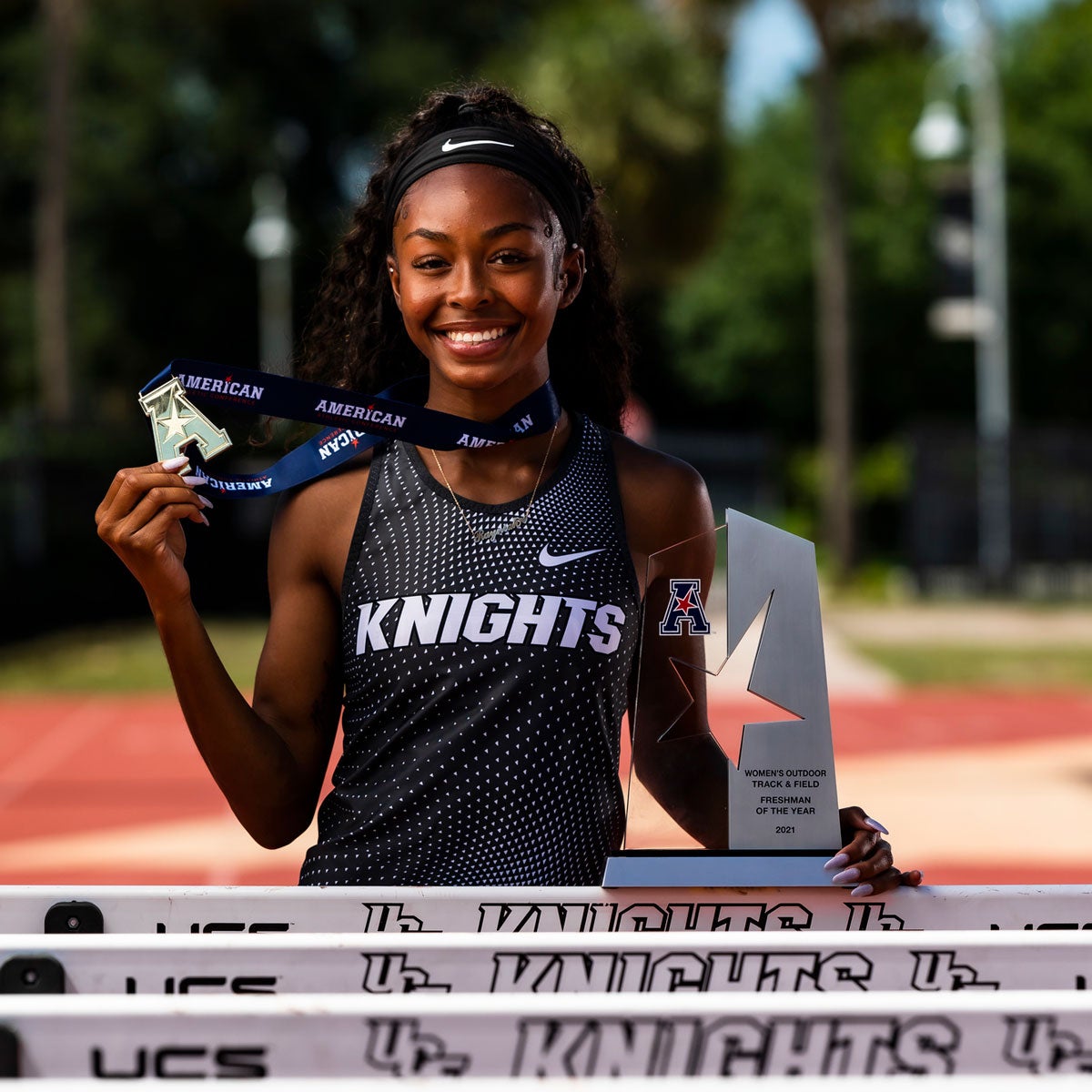 Rayniah Jones stands near hurdles, holding AAC medal and trophy