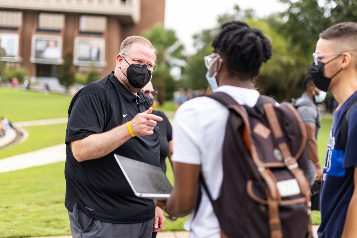 Alex Cartwright wearing a black face mask talks to two male students near Reflecting Pond
