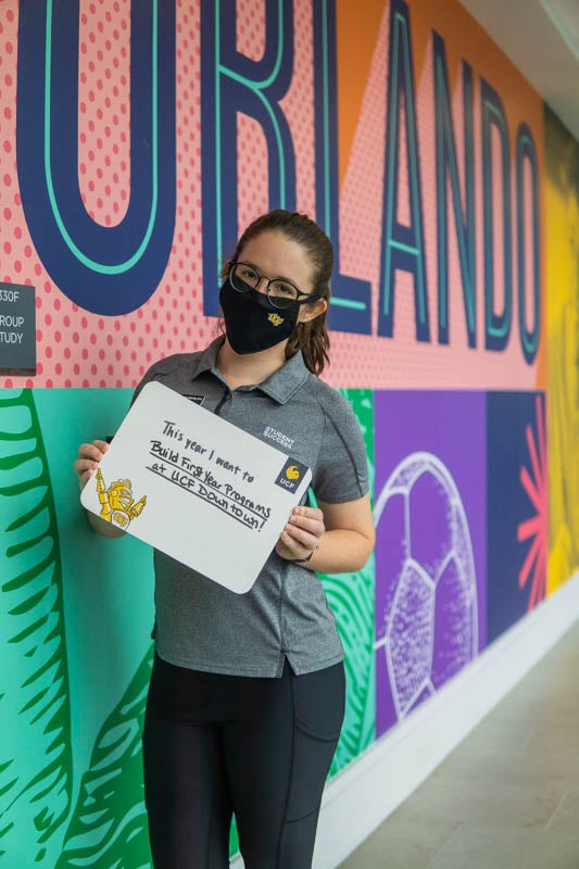 Student wearing gray polo, black pants and UCF mask stands in front of wall mural 