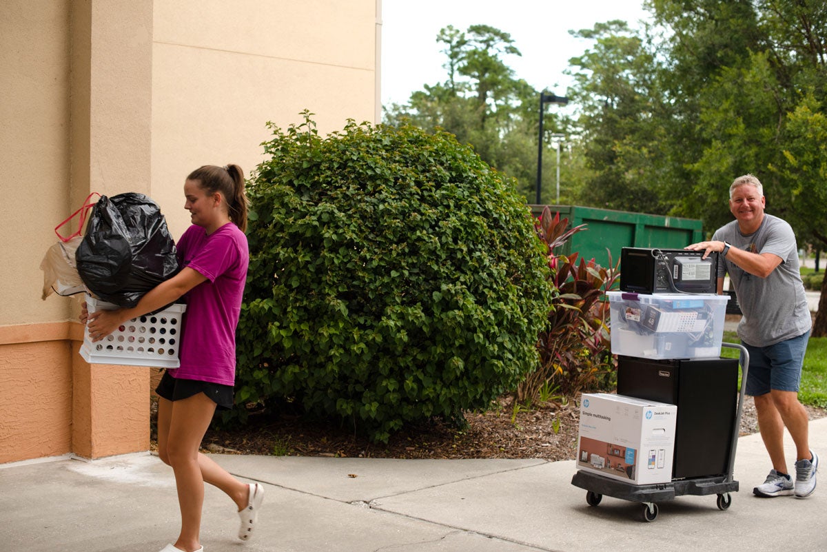 A woman in a pink shirt carries a laundry basket packed with belongings while her father wheels a cart of belongings behind her