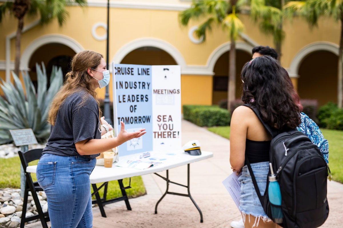 A student talks to two other students at a table with a board promoting the Cruise Line Industry Leaders of Tomorrow