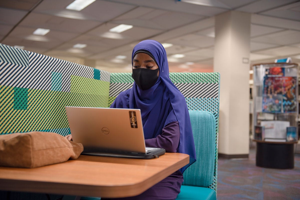 Student wearing purple hijab sits at a table with a laptop in John C. Hitt Library