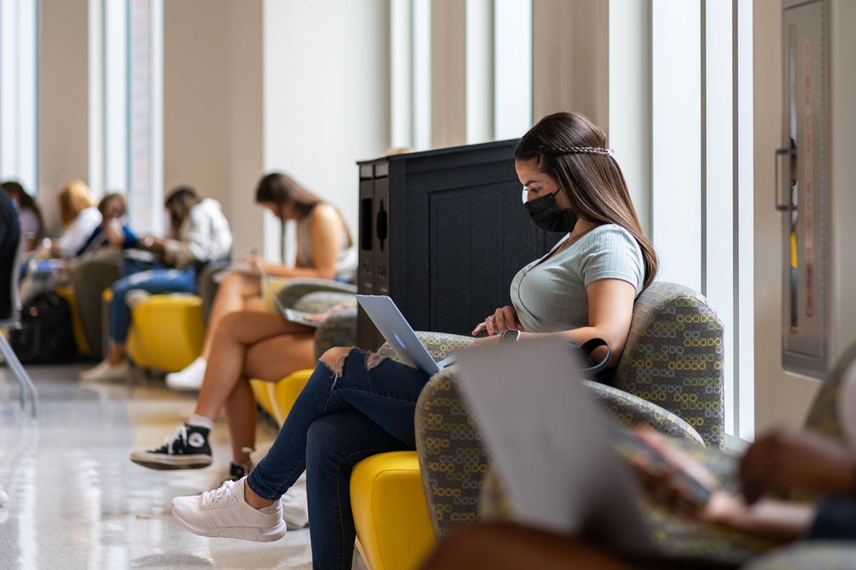 Students wearing masks sit individually in yellow chairs lining a hallway in student union