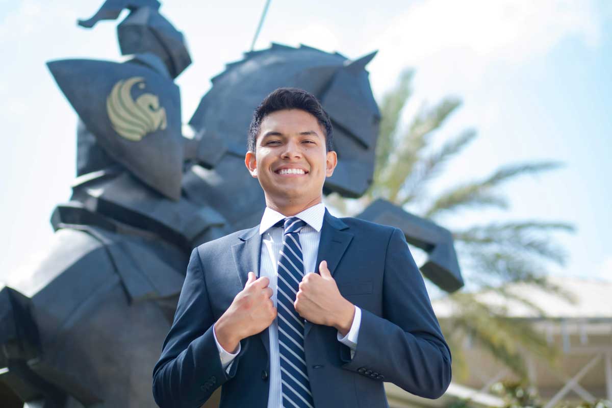 Gonzalo Sauri in a suit stands in front of Victory Knight statue in front of Alumni Center