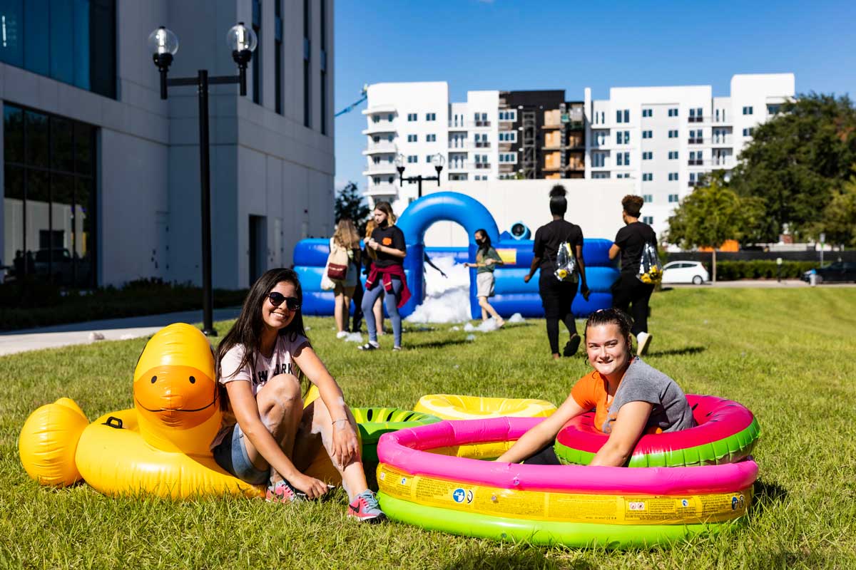 Two women sit in colorful pool floats on grass on sunny day with a blue inflatable rink behind them filled with foam