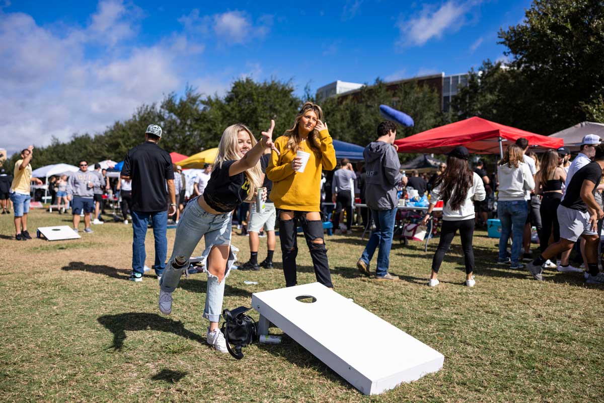 Two female students play cornhole on sunny day near tailgate tents