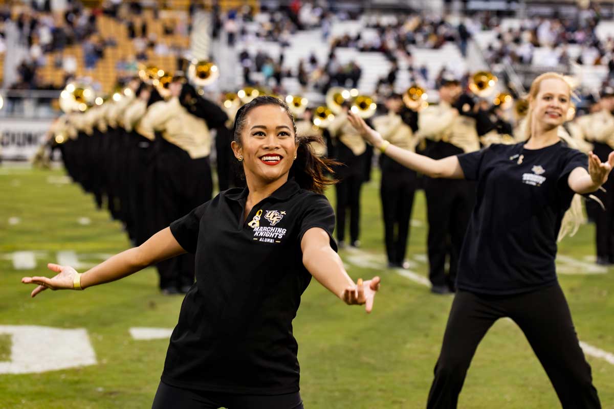 Two alumni Marching Knights dance with horn section playing behind them on the field