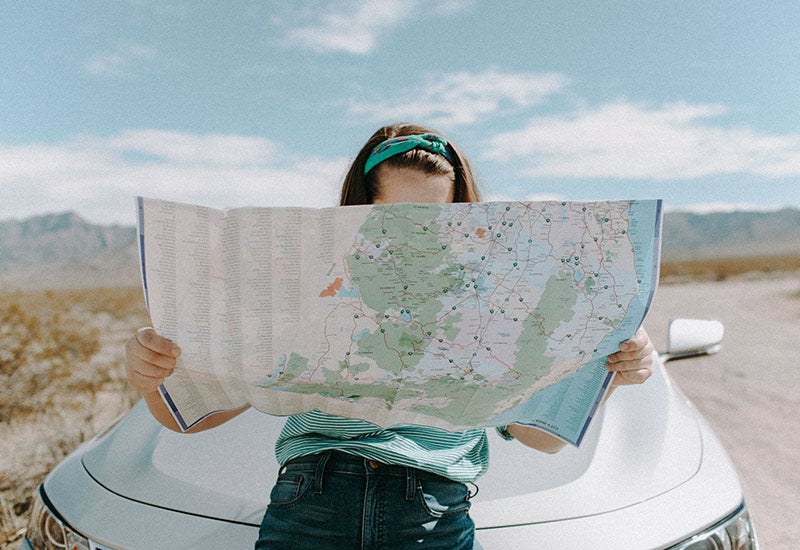 Woman sitting on the hood of her car looking at a paper map.