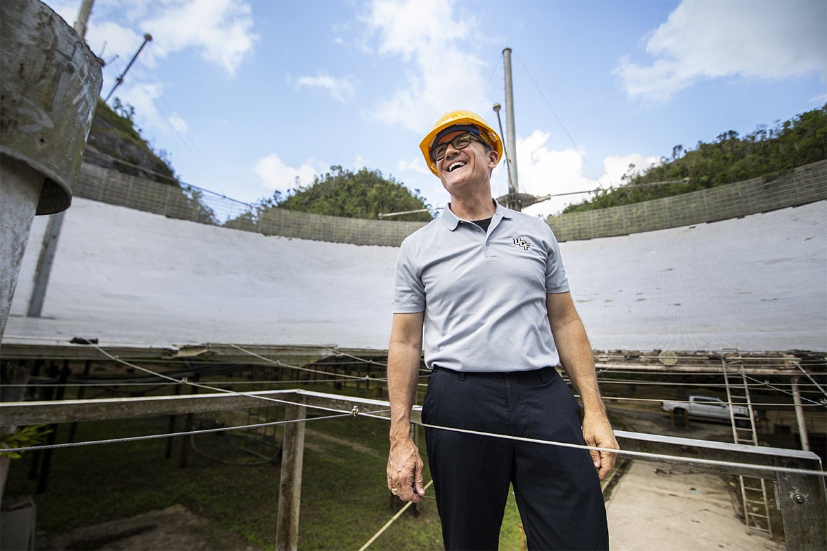 UCF President Dale Whittaker wearing yellow hard hat and glasses, grey polo, black pants, smiling near Arecibo. UCF President Dale Whittaker wearing yellow hard hat and glasses, grey polo, black pants, smiling near Arecibo.