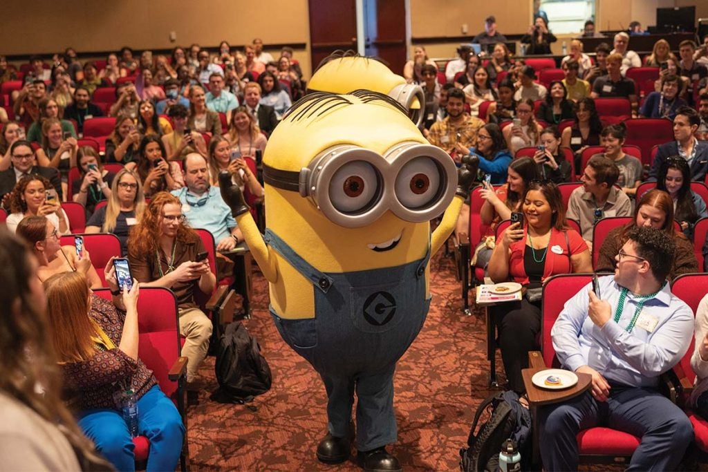 Minion character from Despicable Me walks down the aisle of a crowded auditorium.