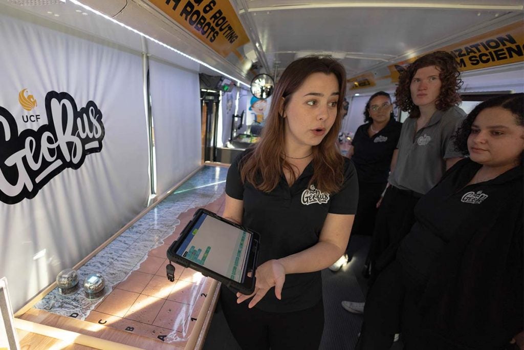 A female UCF student wearing a black collared shirt and black pants is standing onboard the GeoBus, holding an iPad and explaining the Sphero Ball activity to three UCF students.