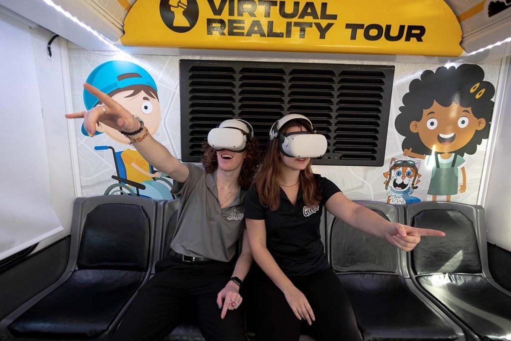 Two UCF students are wearing white virtual reality headsets and are pointing with their index finger into the air while sitting on two of the five black seats in the back of the GeoBus.