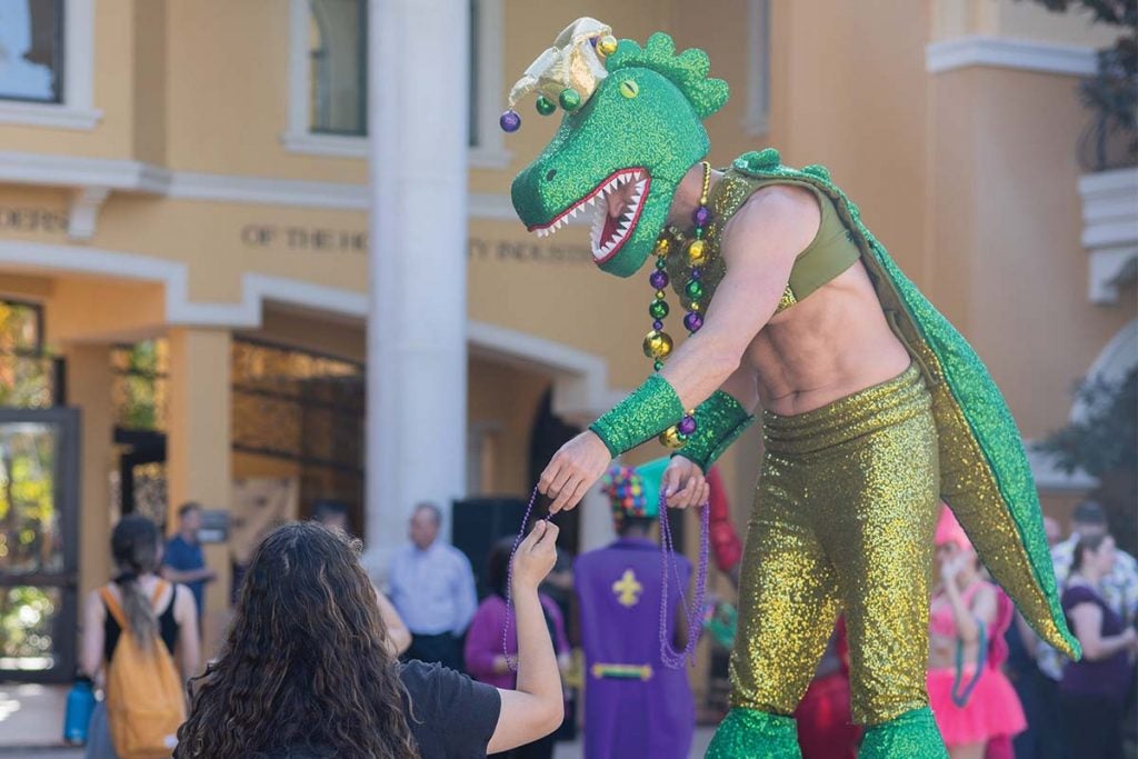 A person dressed up in Mardi Gras themed clothing and a dinosaur masks wears stilts whole giving beads to a student