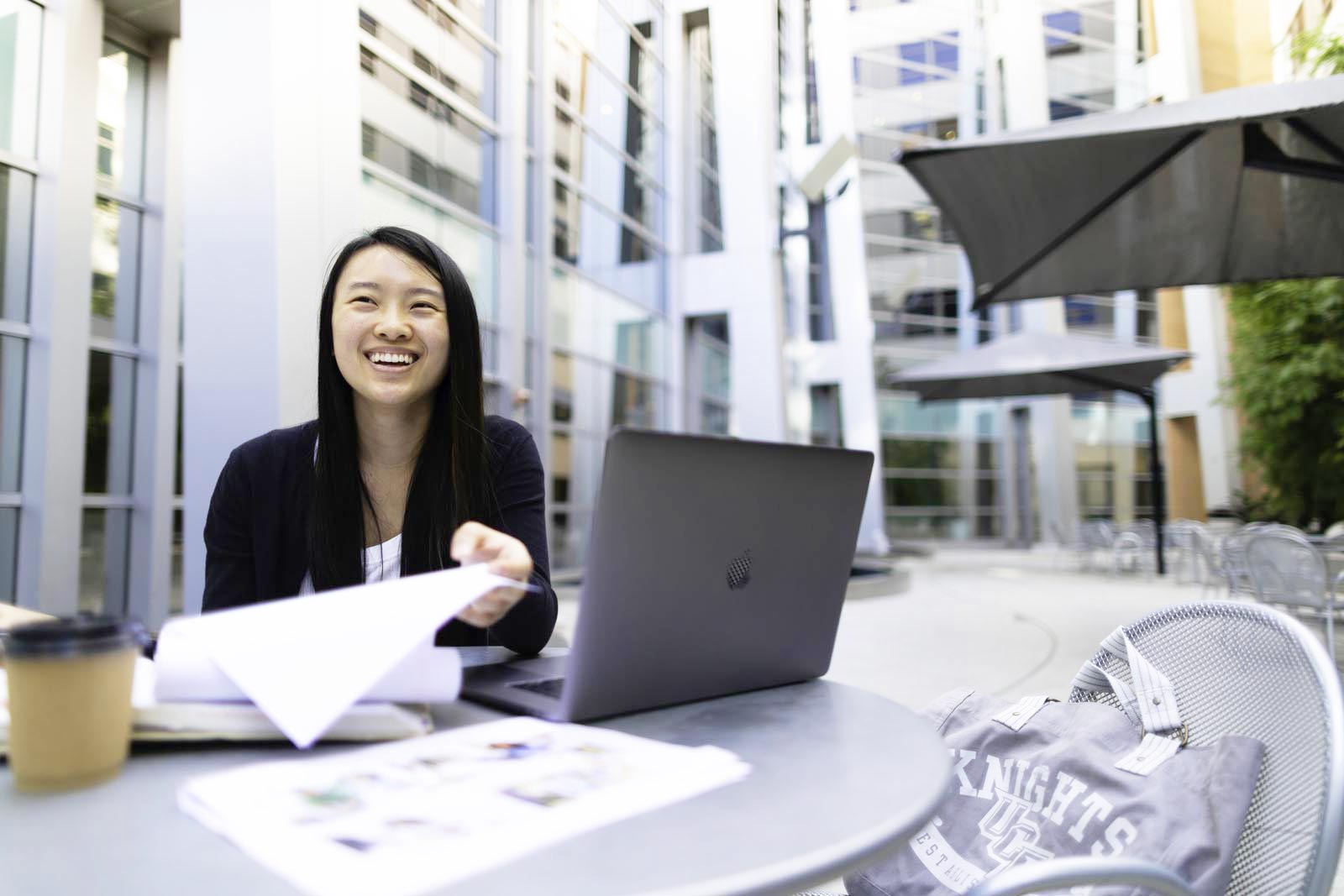 Female student at a courtyard table with laptop and class notes. 