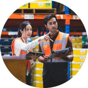 Two warehouse workers in safety vests, holding tablets discussing inventory