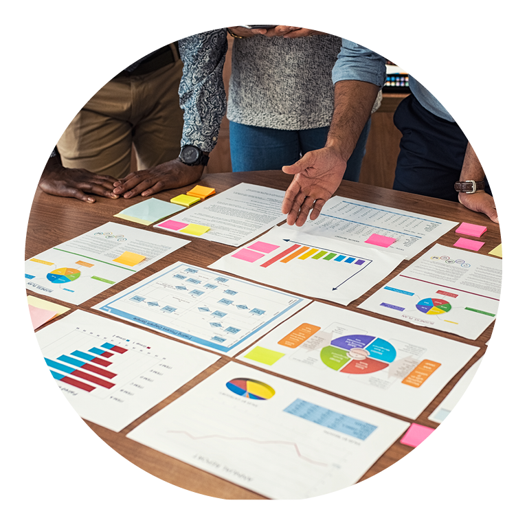 Documents of charts laid out on a table with several people discussing them.