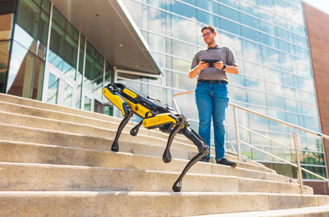 A student from the Robotics Club of Central Florida at UCF uses a handheld controller to operate a four-legged robotic dog on the stairs outside the library.