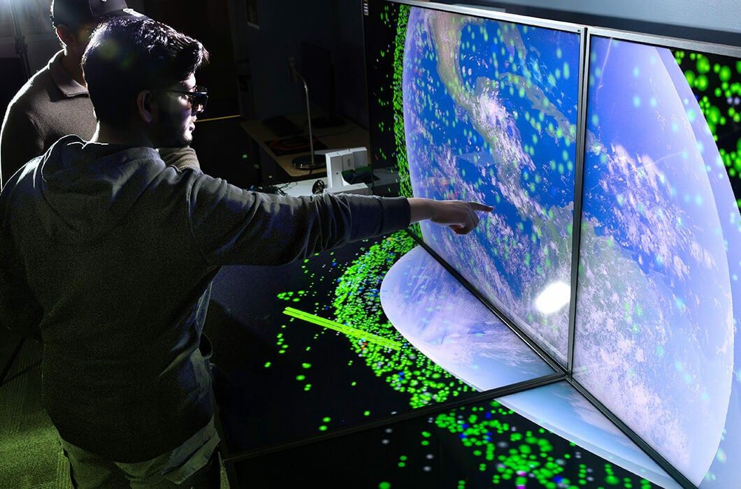 Two students stand in front of large computer screens displaying a map of the world as they learn through simulation technology in the Virtual and Augmented Reality Lab at UCF’s Institute for Simulation and Training.
