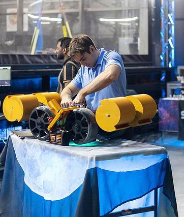 A man in a blue shirt works on a yellow and black robotic device from Exolith Lab, placed on a table covered with a blue and white cloth in an indoor setting.