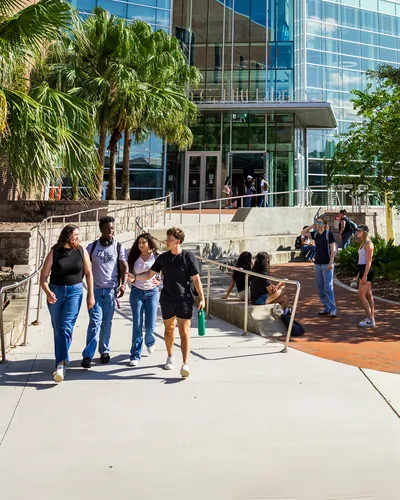 Four University of Central Florida students walk toward the camera on a wide campus walkway while others sit along a low wall; a glass-front UCF library entrance and palm trees fill the background in bright afternoon sun.