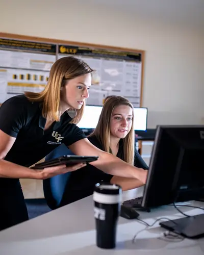 UCF students review code together as a mentor in a UCF polo points at a desktop screen in a campus lab.