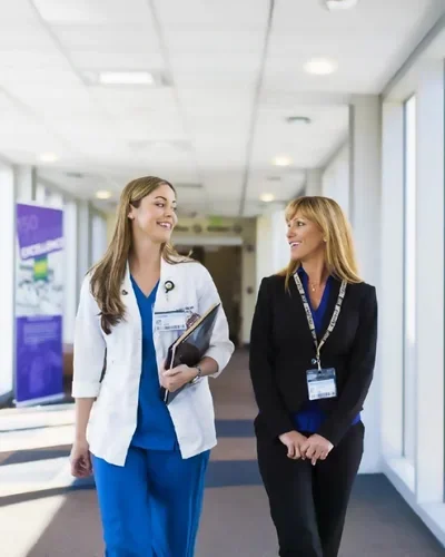 UCF nursing student in a white coat and blue scrubs walks with a clinical mentor down a bright hospital corridor.