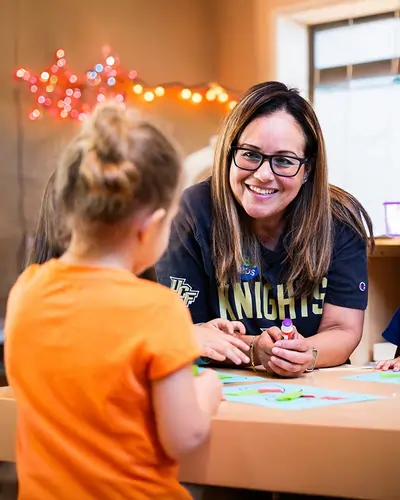 UCF educator smiles and helps a young child with an arts-and-crafts activity at a classroom table.