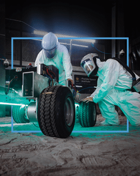 Two people in protective suits work on a rover in the Exolith Lab