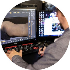 Person using a stylus at dual monitors displaying a 3D model of an arm.