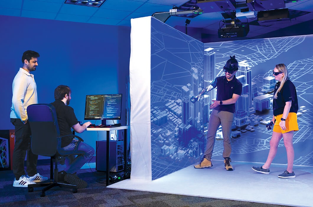 Four people interact in a tech lab; one codes at a computer while three try out virtual reality gear in front of a digital cityscape projection.