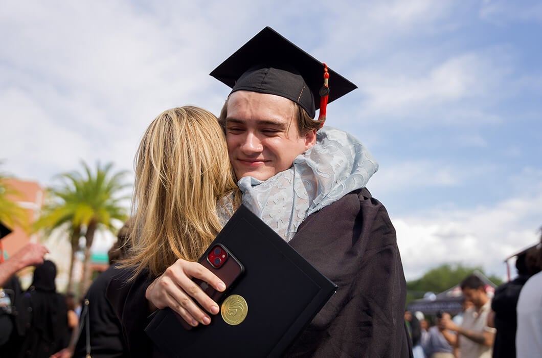 A young graduate in a cap and gown smiles while hugging a woman outdoors. He holds a diploma and a phone, and there are palm trees and other people in the background under a partly cloudy sky.