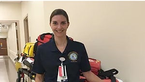 Smiling nurse standing in a hallway with medical gear behind her.