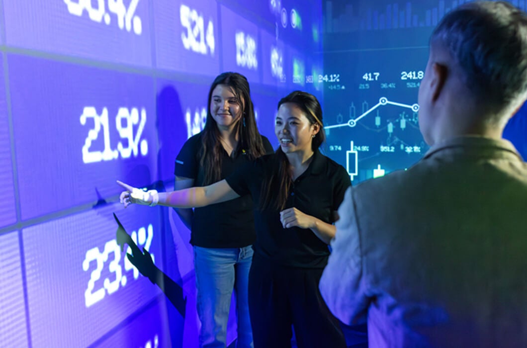 Three people stand in front of a large digital screen displaying charts and percentages. One woman points at the data while others watch, suggesting a discussion or presentation about financial or statistical information.