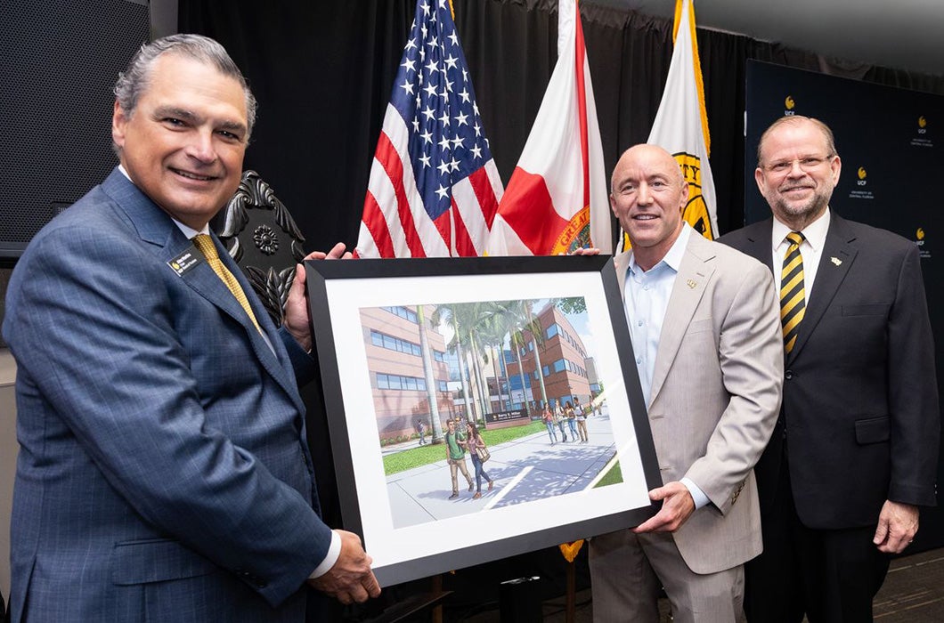Alex Martins, Alexander Cartwright and Barry Miller pose together, smiling, as two of them hold a framed architectural rendering of a the UCF College of Business building. Behind them are American and Florida flags and a UCF (University of Central Florida) flag.