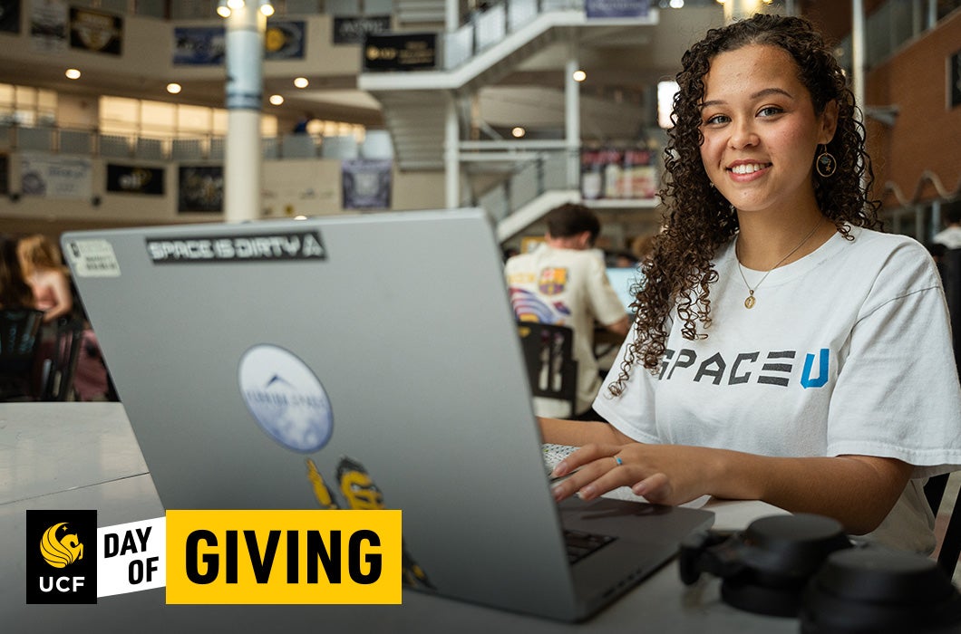 A young woman smiles while working on a laptop covered in stickers. She wears a SPACE U shirt. The setting is a bright, modern indoor space. A UCF Day of Giving logo appears in the corner.