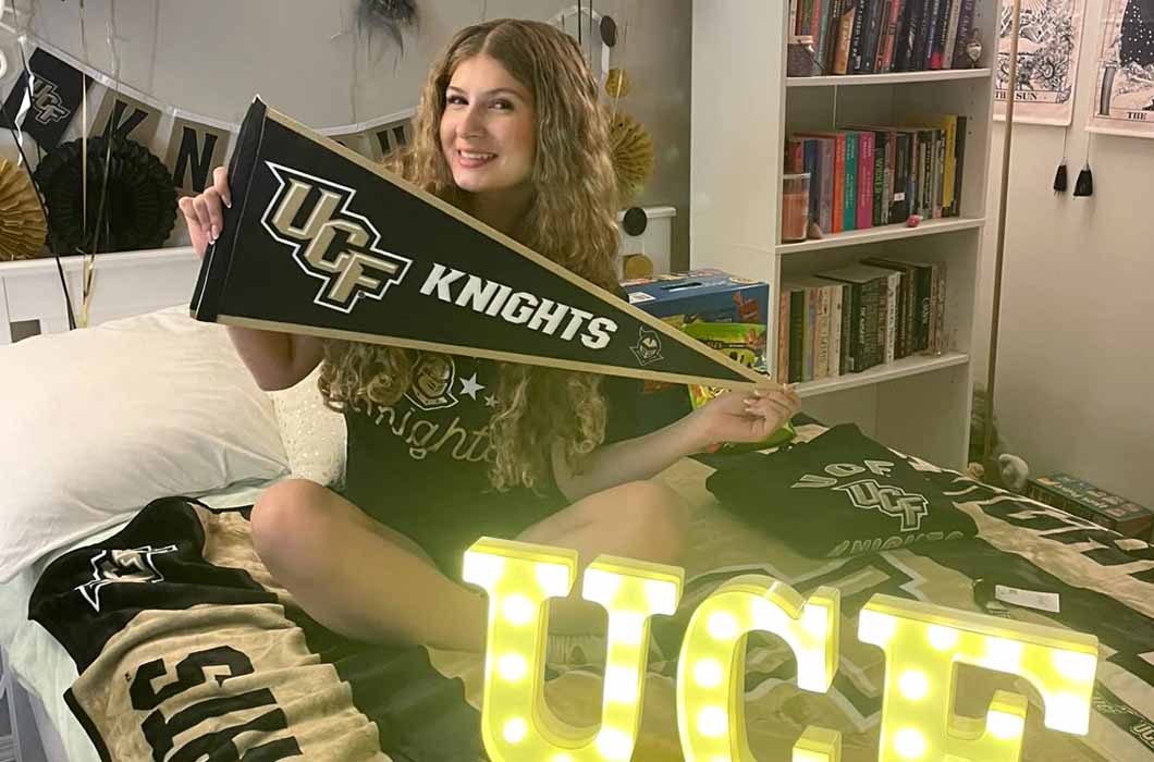 A young woman sits on a bed holding a UCF Knights pennant, surrounded by University of Central Florida merchandise. The bed has UCF-themed blankets and a large illuminated UCF sign in front of her.
