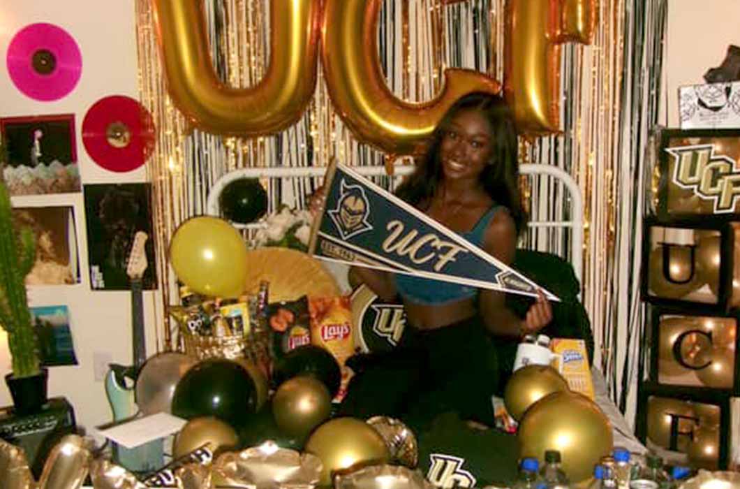 A woman sits on a bed decorated with gold and black balloons, streamers, and UCF signs. She holds a UCF pennant and smiles. The room features snacks, party items, and UCF-themed decor. " width="1060" height="700" class="alignnone size-full wp-image-94076 img-fluid