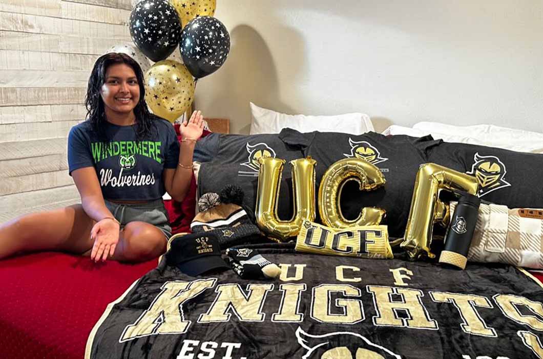 A smiling young woman sits on a bed covered in UCF Knights gear, including a blanket, beanie, tumbler, and gold UCF balloons. She wears a “Windermere Wolverines” shirt and raises her hands. Black and gold balloons are in the background. 