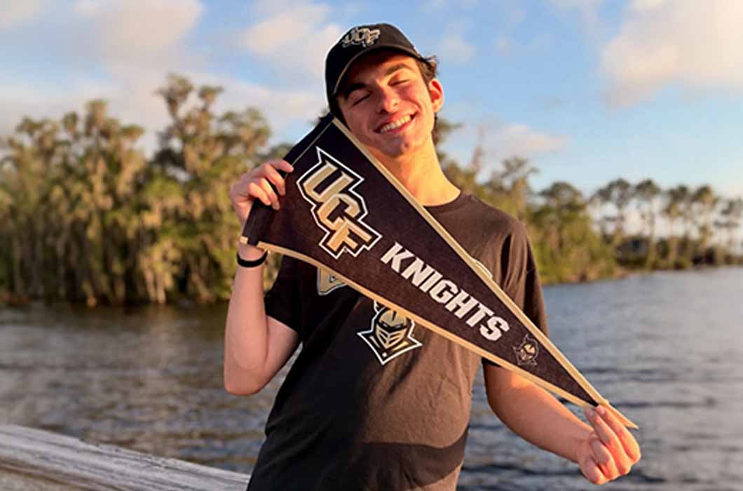 A smiling person wearing a UCF hat and shirt holds a UCF Knights pennant while standing outdoors near water with trees in the background. 