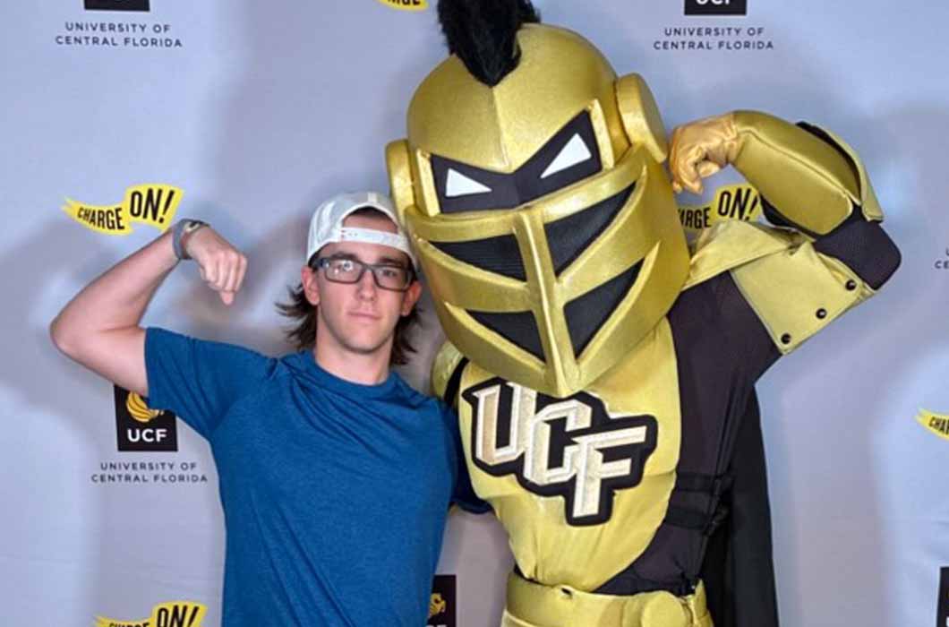 A person in a blue shirt and white cap poses, flexing their arm, next to UCF’s knight mascot in gold armor at a University of Central Florida event backdrop.